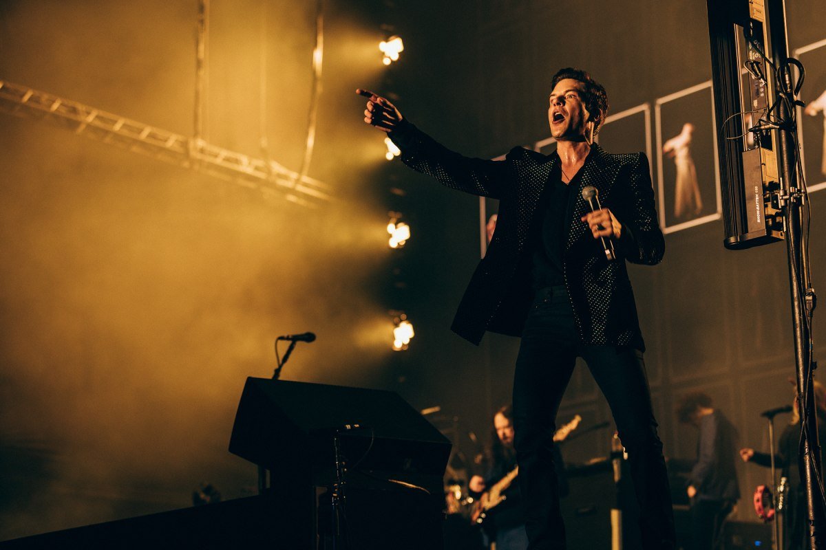 Brandon Flowers, vocalista da banda The Killers, canta no palco segurando um microfone e apontando para a plateia. Ao fundo, uma luz amarela - Metrópoles