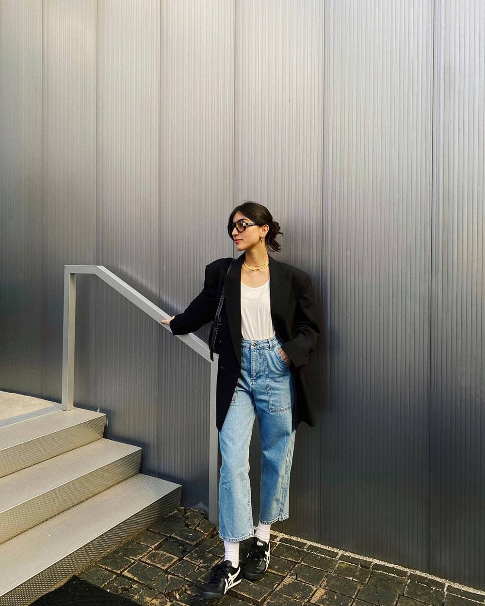 A influenciadora digital Maju Trindade, uma mulher jovem, branca, com cabelo liso longo, posando para foto em uma escada cinza. Ela veste uma blusa branca, calça jeans clara, blazer e tênis, ambos pretos, e óculos de sol. - Metrópoles