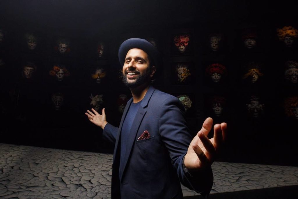 Homem de roupa preta e chapéu olhando para a foto sorrindo e de braços abertos