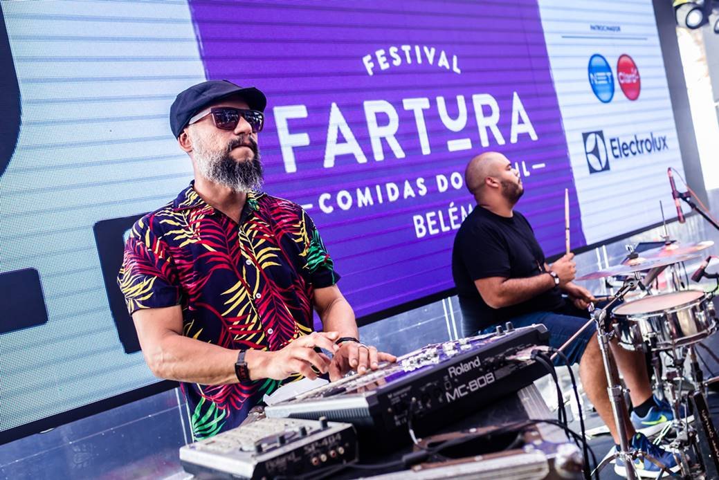Foto de um homem mixando mesa de DJ ao lado de um outro homem que toca bateria. Ao fundo, um telão de led escrito Festival Fartura
