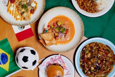 Fotos de uma mesa com vários pratos em cima e comidas diferentes, também tem uma bandeira do Brasil e uma pequena bola de futebol e cima da mesa