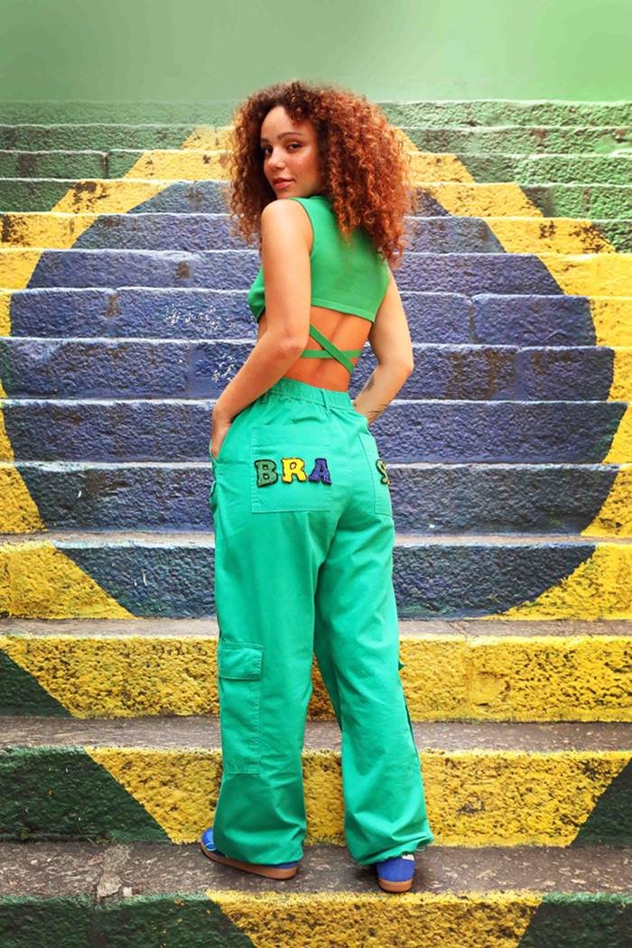 Mulher jovem e negra, de cabelo cacheado castanho, posando para foto em uma espada pintada com a bandeira do Brasil. Ela usa um top verde com amarrações, uma calça verde clara folgada com as letras B R A bordadas e tênis azul.