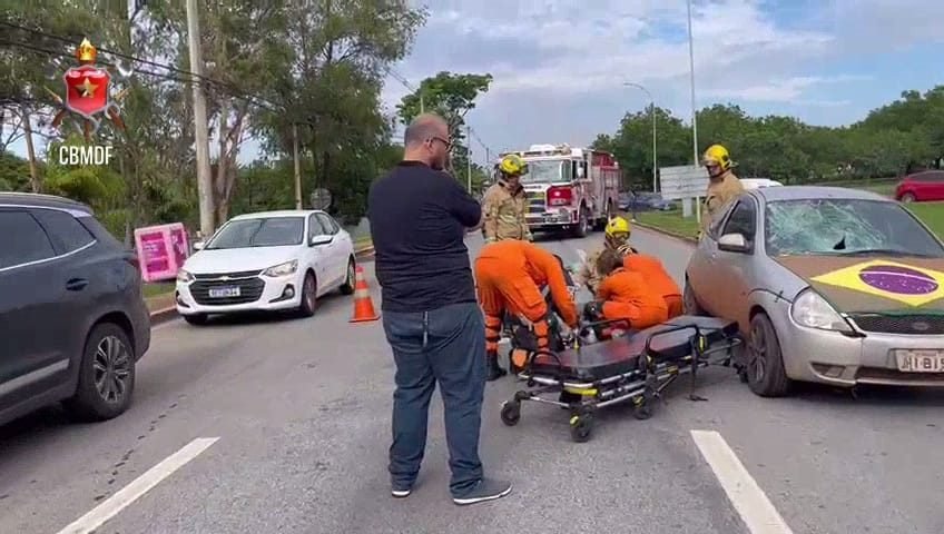 Bombeiros em uniforme laranja colocam homem em maca. Pedestre olha atendimento. Foto colorida
