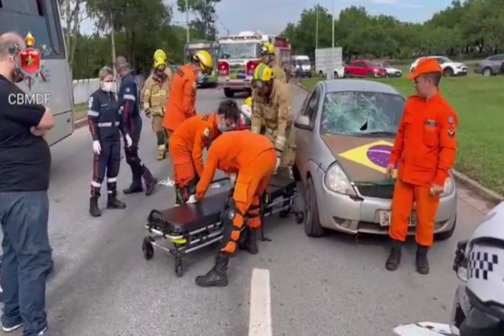 Bombeiros em uniforme laranja colocam homem em maca. Pedestre olha atendimento. Foto colorida