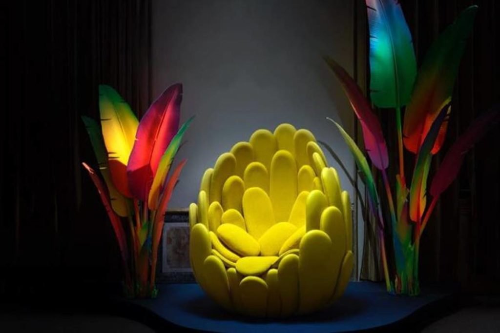 cadeira amarela parecida com flor amarela e folhas de palmeiras coloridas dos lados - metrópoles