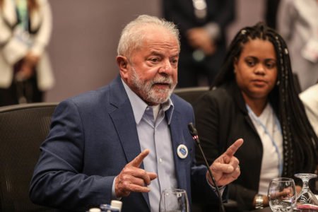 O presidente eleito Lula discursa na COP27, no Egito. Ele aparece sentado diante de microfone, gesticulando, ao lado de mulher negra - Metrópoles