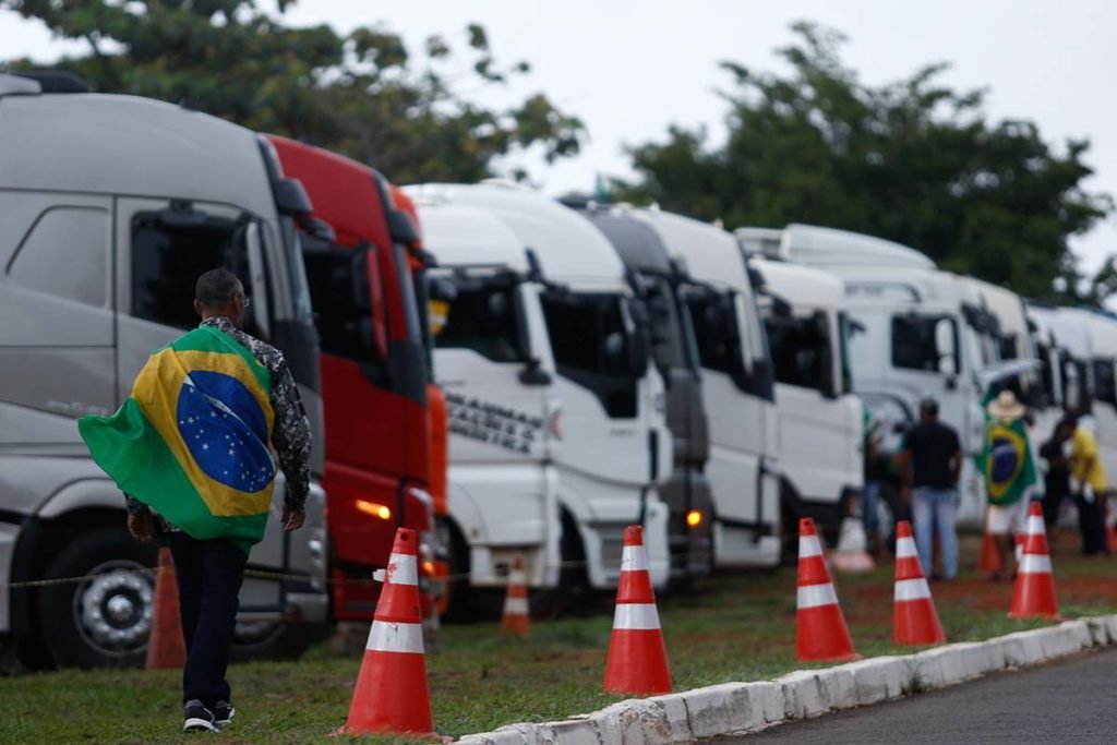 Homem usando bandeira do Brasil como capa anda próximo a caminhões estacionados. Nota das Forças Armadas, que critica