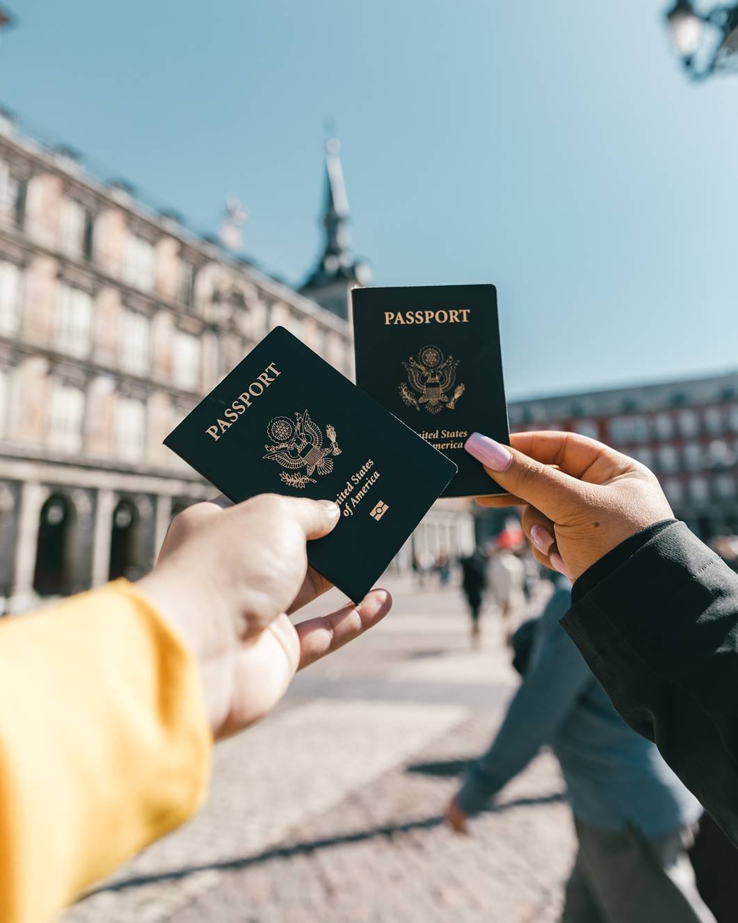 Foto mostra duas mãos segurando um passaporte cada uma, ao fundo mostra uma cidade famosa por seu turismo