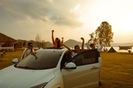 Foto de um carro parado durante um por do sol, cinco pessoas estão ao lado do carro para as mãos para cima