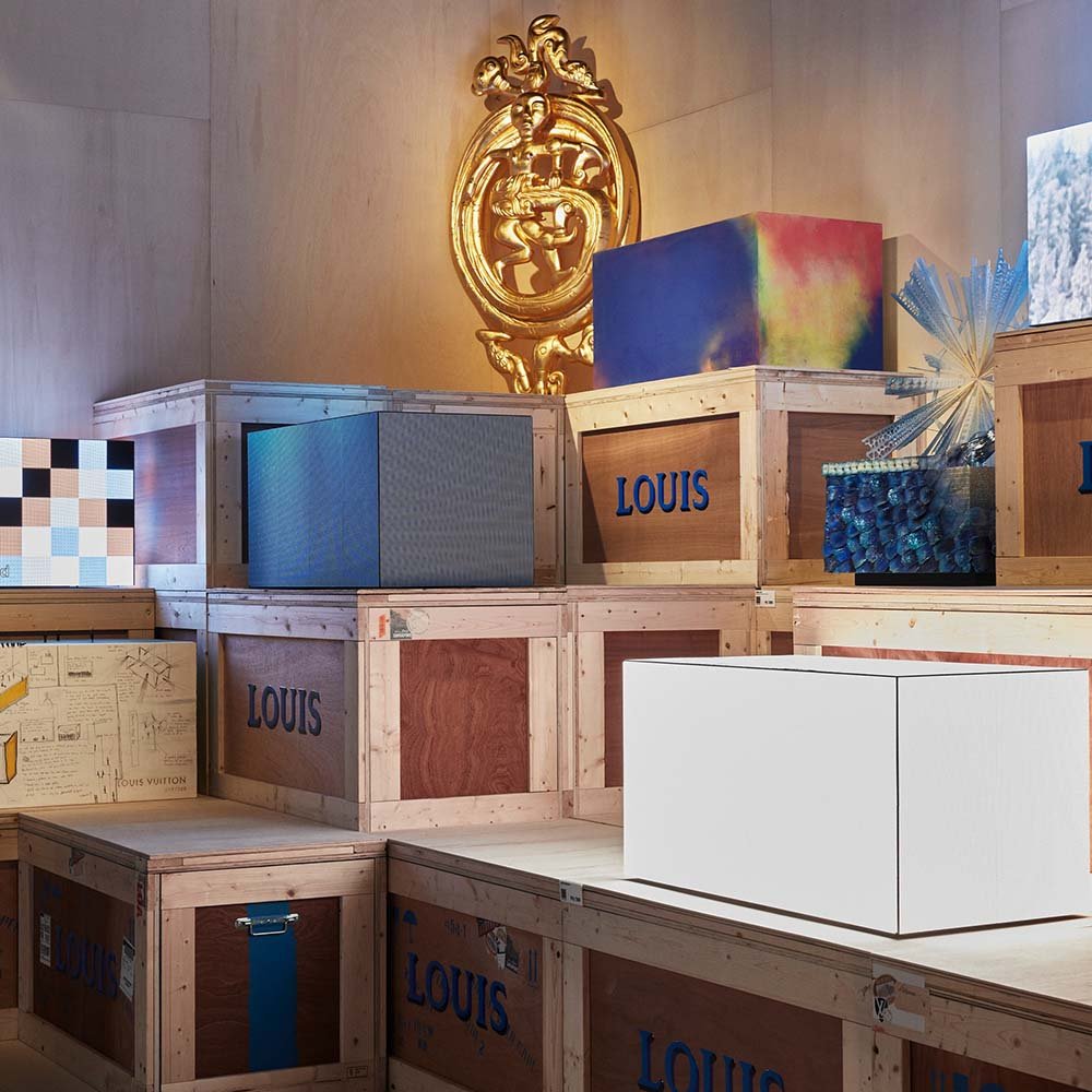Exposição da marca Louis Vuitton em Nova York. Na foto é possível ver vários caixotes de madeira empilhados. Eles possuem a logomarca da grife pintada de azul.