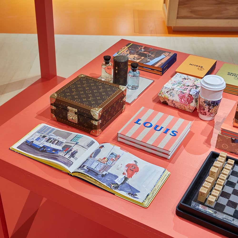 Exposição da marca Louis Vuitton em Nova York. Na foto é possível ver uma mesa de madeira vermelha com vários objetos em cima, como uma pequena caixa com a logomarca da grife francesa, livros sobre a Louis Vuitton e outros objetos de decoração da empresa.