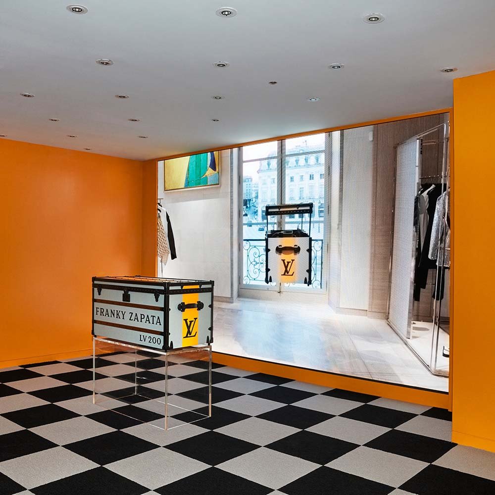 Exposição da marca Louis Vuitton em Nova York. Na foto é possível ver um quarto com paredes laranjas, um chão com cerâmica quadriculada preto e branco e, no centro, um baú, também preto e branco, com a logo marca da Louis Vuitton e o nome Franky Zapata bordado