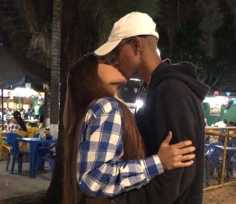 Menina com cabelo liso grande e blusa xadrez beija menino com boné branco e casaco preto