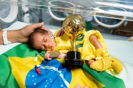 Foto de um bebê de amarelo deitado em cima de uma bandeira com um trófeu dourado na frente