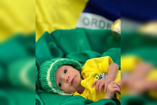 Foto de um bebê de verde e amarelo e com fundo da bandeira atrás