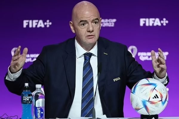 Foto de um homem careca e de terno na frente de um telão da Fifa falando com os braços abertos sobre a Copa do Mundo