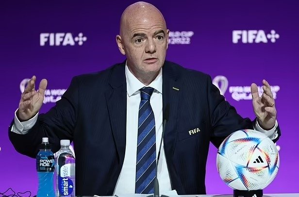 Foto de um homem careca e de terno na frente de um telão da Fifa falando com os braços abertos