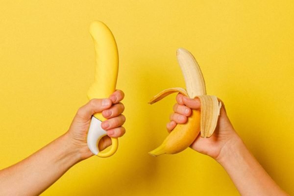 Mãos segurando um sex toy e uma banana com fundo amarelo - Metrópoles