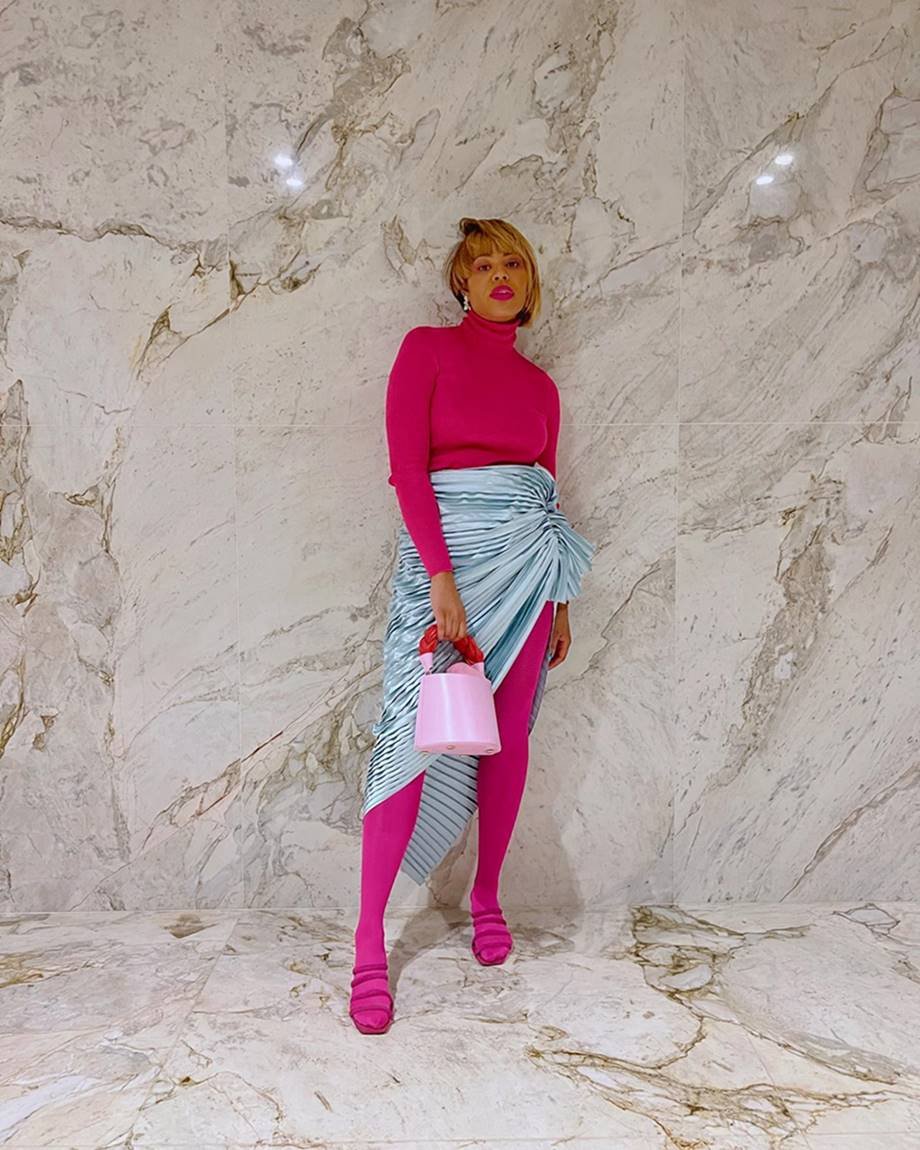 A influencer Larissa Cunegundes, uma mulher jovem, negra, de cabelo liso curto, posando para foto em um parede de mármore. Ela usa uma blusa de manga comprida rosa, meia calça rosa, uma saia prata metalizada e uma sandália de salto também rosa