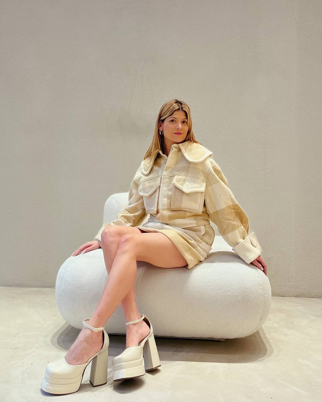 A jornalista Giovana Romani, uma mulher branca, jovem e com cabelo liso loiro, posando para foto em um sofá bege. Ela usa um conjunto de blusa e saia xadrez, em tons claros, e uma sandália plataforma branca