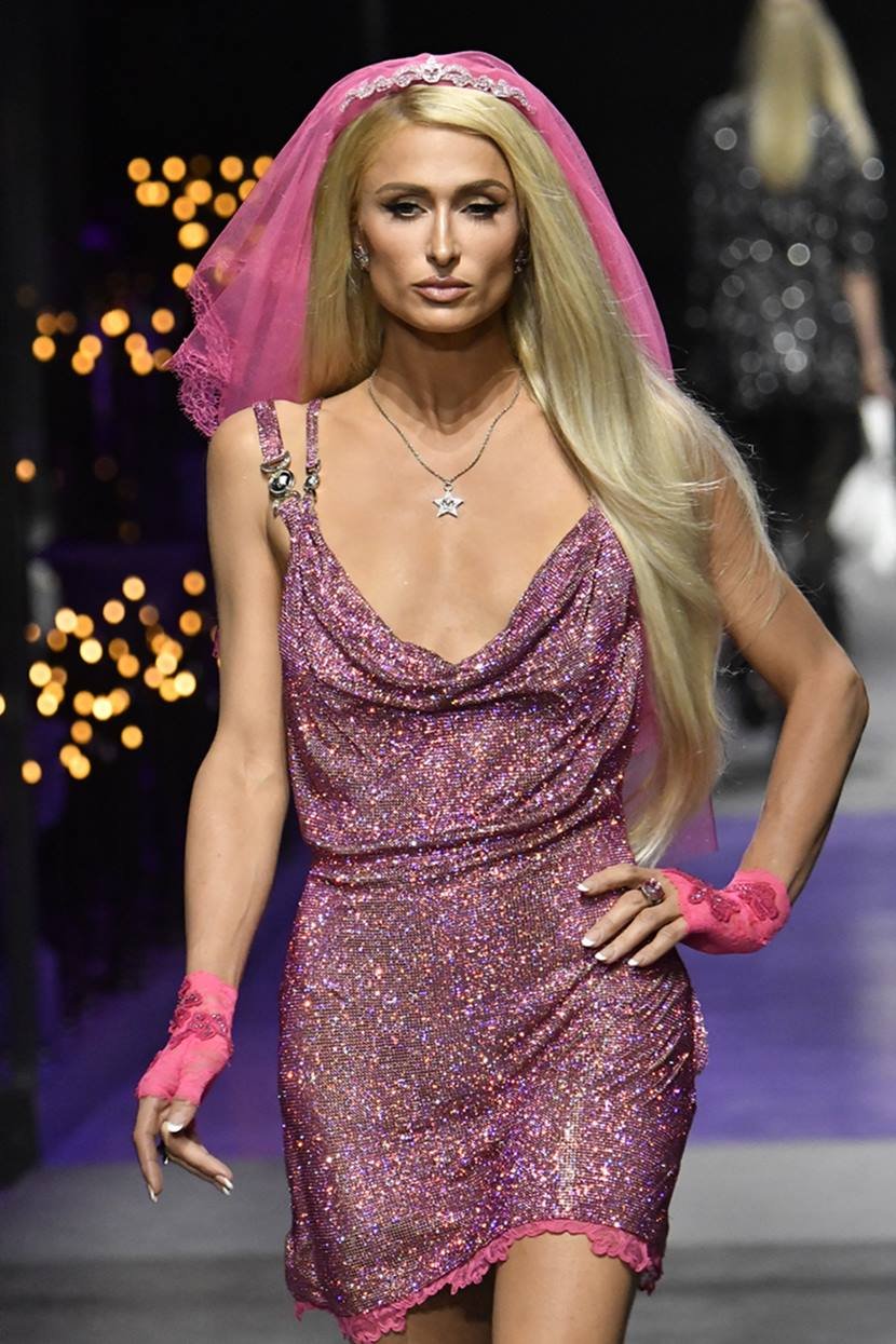 A socialite e empresária Paris Hilton desfilando na passarela da marca Versace na Semana de Moda de Milão. Ela usa um vestido curto brilhante e um véu rosa de tule preso à cabeça. - Metrópoles