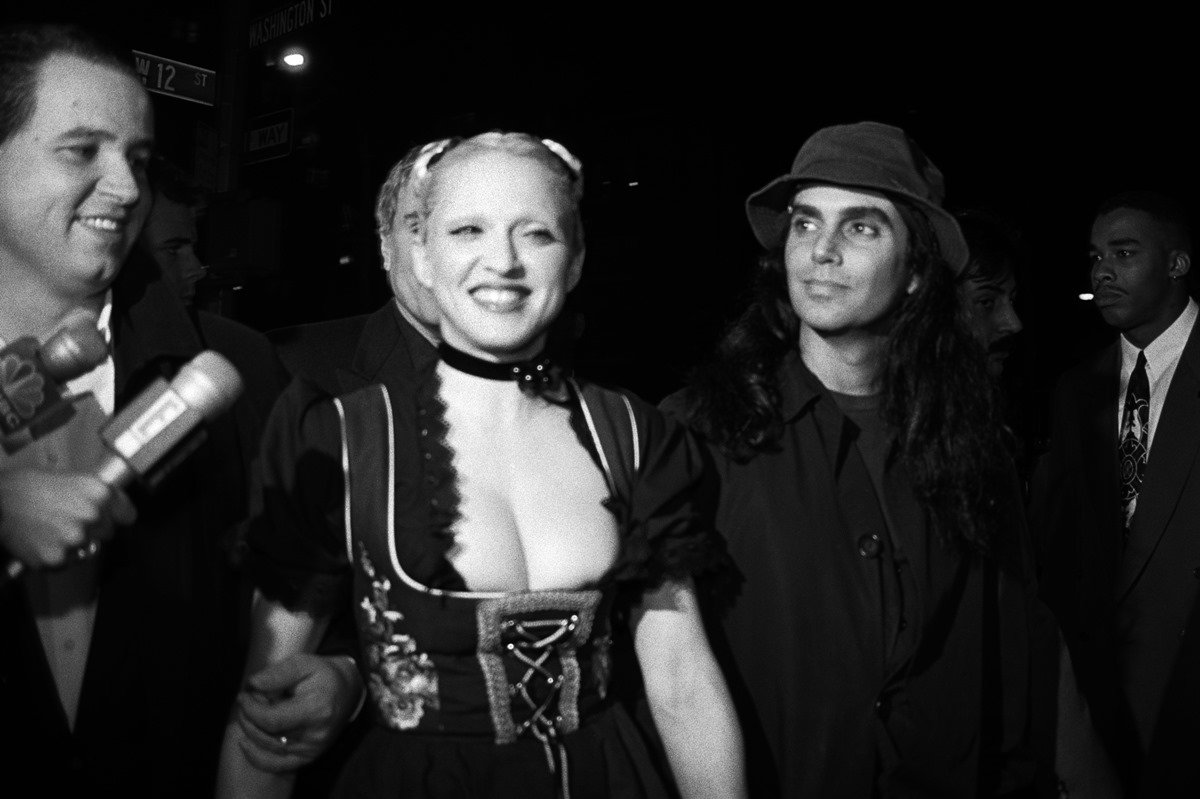 Madonna e o fotógrafo Steven Meisel chegam à festa de publicação do livro "Sex" de Madonna em 15 de outubro de 1992 no Industria Superstudio em Nova York, Nova York. Ambos são jovens, mas ela é uma mulher branca de cabelo liro. Ele tem a pele morena e o cabelo liso longo.