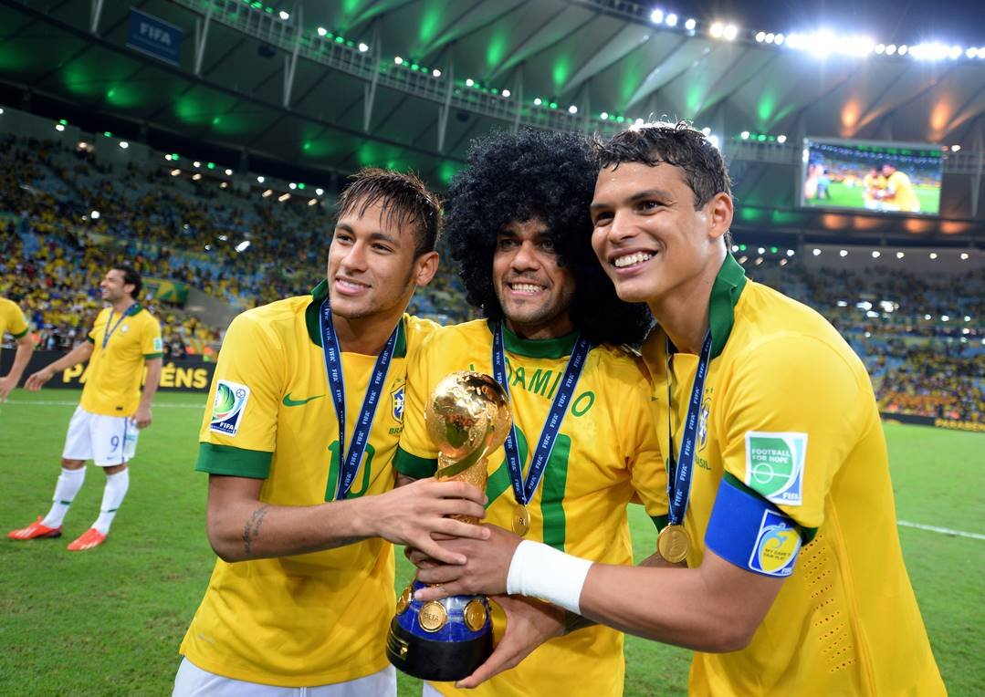 Foto de Neymar, Daniel Alves e Thiago Silva comemorando a final das Copas das Confederações. Os três usam a camisa amarela do Brasil e seguram a taça da disputa. No meio, Daniel Alves usa uma peruca e os três estão usando uma medalhe