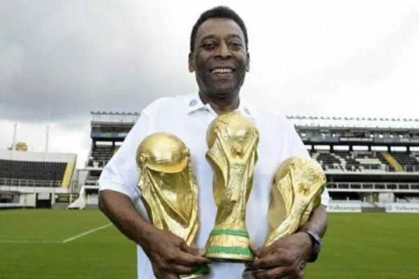 Pelé, ex-jogador de futebol e da seleção, um dos maiores jogadores do Brasil e do mundo, segura três taças da Copa do Mundo sorrindo em campo de um estádio - Metrópoles