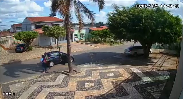 Imagem de câmera de segurança mostra carro preto em rua. Um homem desce e aborda um pedestre com arma de fogo. Foto colorida