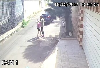 Imagem de câmera de segurança mostra carro preto em rua. Um homem desce e aborda um pedestre com arma de fogo. Foto colorida