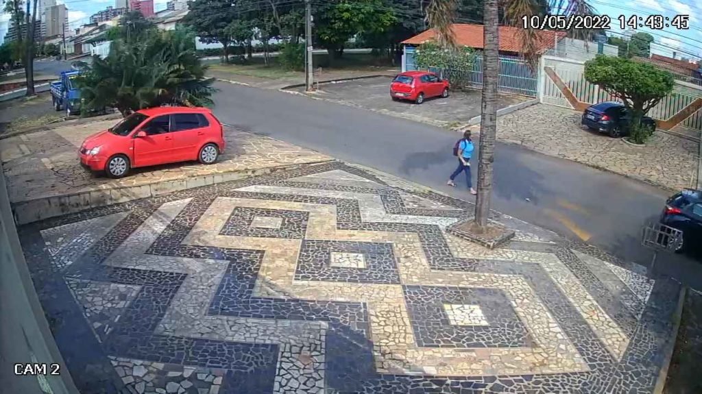 Imagem de câmera de segurança mostra carro preto em rua. Um homem desce e aborda um pedestre com arma de fogo. Foto colorida