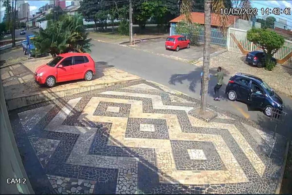 Imagem de câmera de segurança mostra carro preto em rua. Um homem desce e aborda um pedestre com arma de fogo. Foto colorida