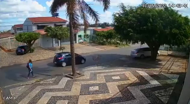 Imagem de câmera de segurança mostra carro preto em rua. Um homem desce e aborda um pedestre com arma de fogo. Foto colorida