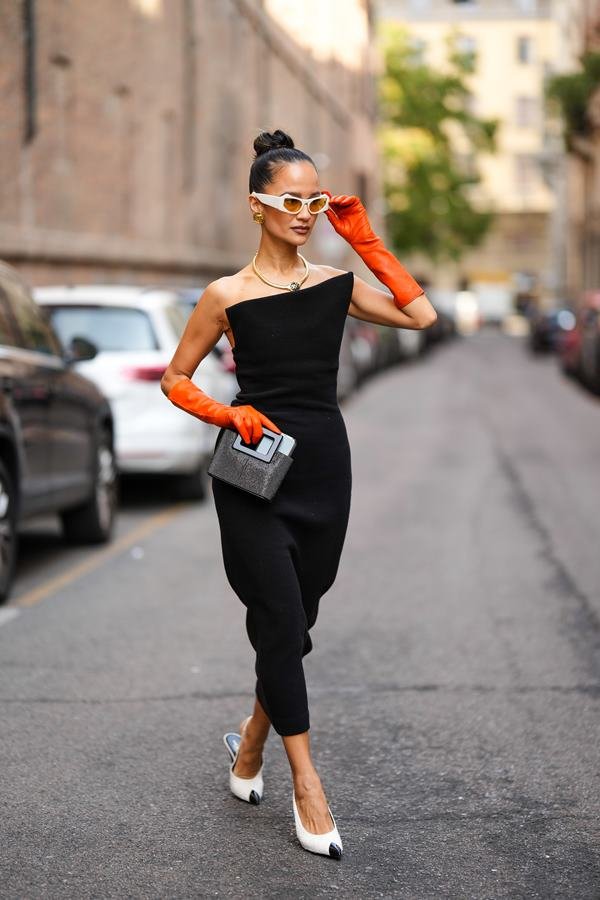 Mulher branca andando na rua. Ela está fazendo pose para foto, usa vestido preto e luvas laranjas