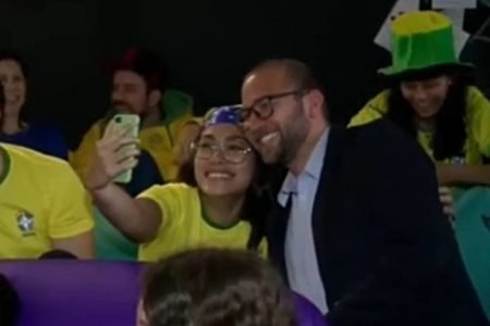 foto de uma mulher com blusa do brasil ao lado de um homem de terno e oculos tirando uma foto - metrópoles