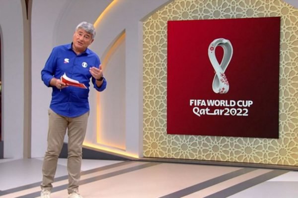 Foto do narrador Cleber Machado, usando uma camisa azul e uma calça bege nos estúdios da Globo. Ao seu lado, um painel temático da Copa do Mundo de 2022 no Catar - Metrópoles