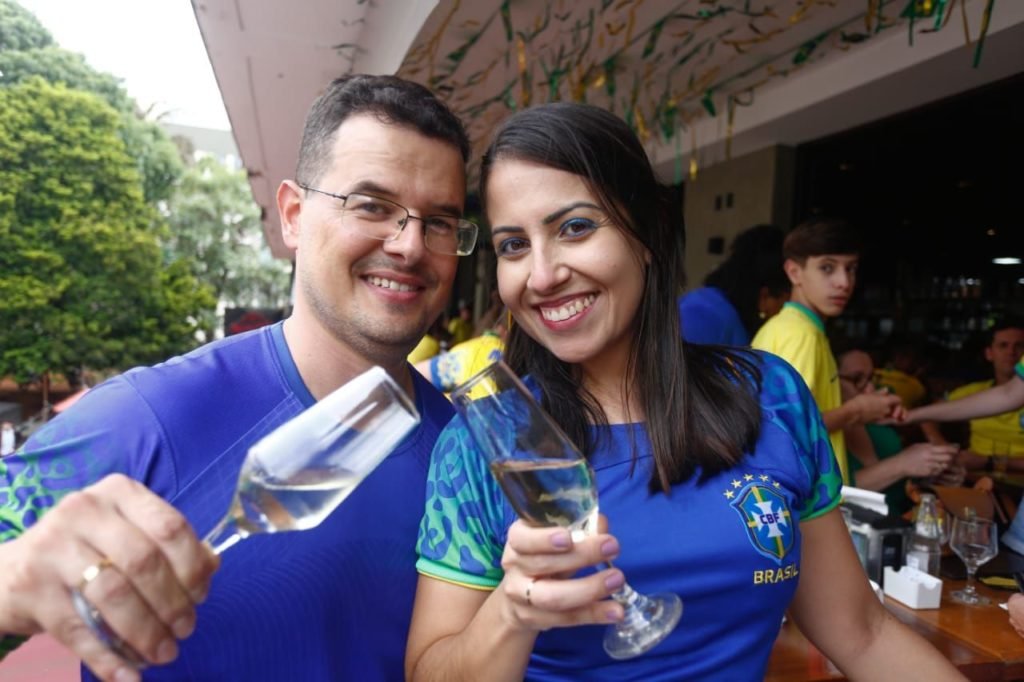 Homem branco de cabelos negros e curtos sorri para foto e segura taça com bebida alcóolica. Ele usa óculos. Mulher de pele branca e cabelos morenos sorri para câmera. Ela segura uma taça com bebida alcóolica e veste uma camisa azul da seleção Brasileira. Foto colorida