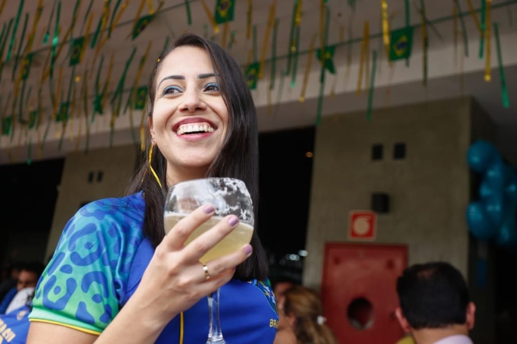 Mulher de pele branca e cabelos morenos sorri para câmera. Ela segura uma taça com bebida alcóolica e veste uma camisa azul da seleção Brasileira. Foto colorida