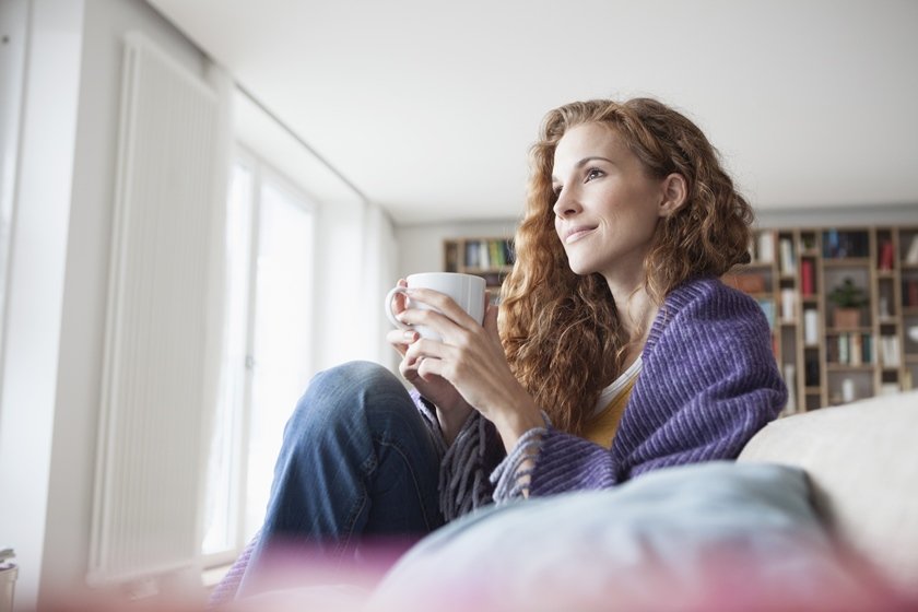 Uma mulher branca de cabelos ondulados está sentada no sofá embrulhada com uma coberta e tomando chá - Metrópoles