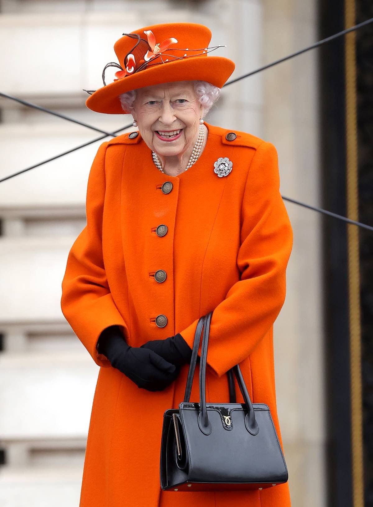 Rainha Elizabeth II posa para foto em com um chápeu e um casaco laranja. Usa ainda luvas pretas e segura, na mão esquerda, uma bolsa de couro preta. - Metrópoles
