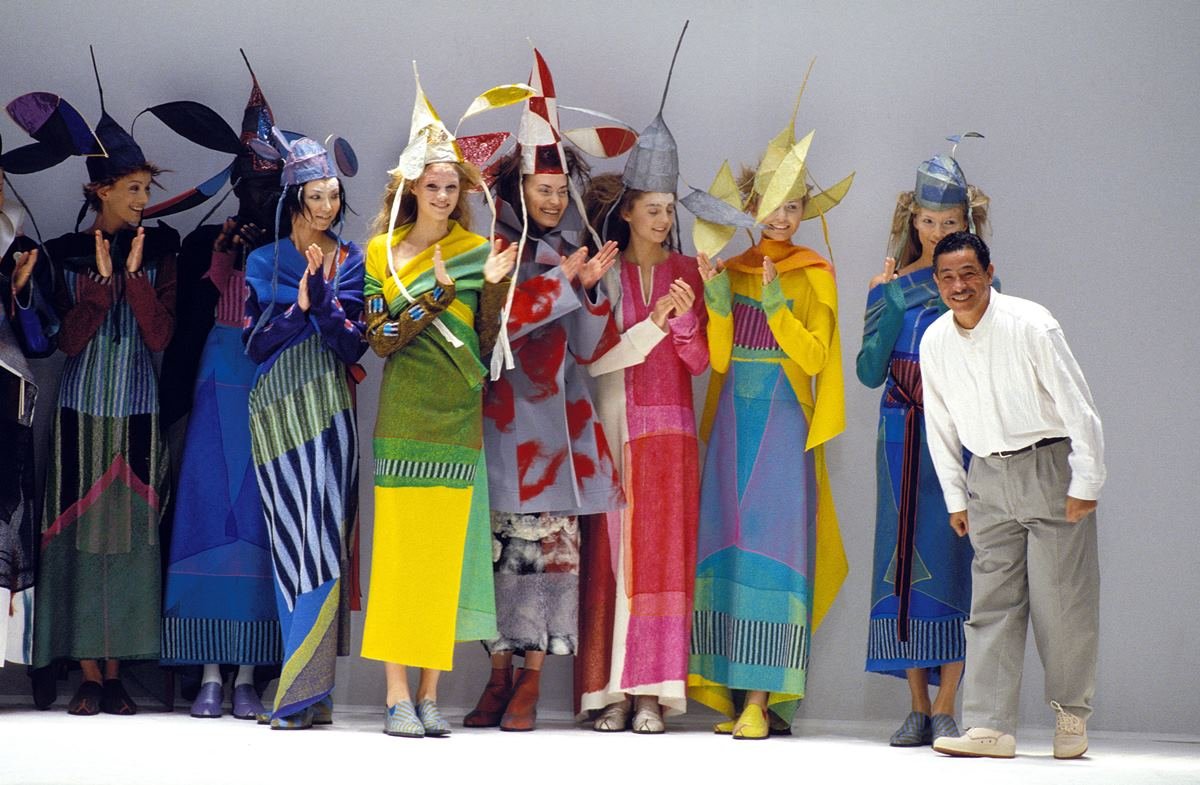O estilista Issey Miyake no final da apresentação de um desfile de 1997. Na foto é possível ver modelos usando roupas e chápeus coloridos. - Metrópoles
