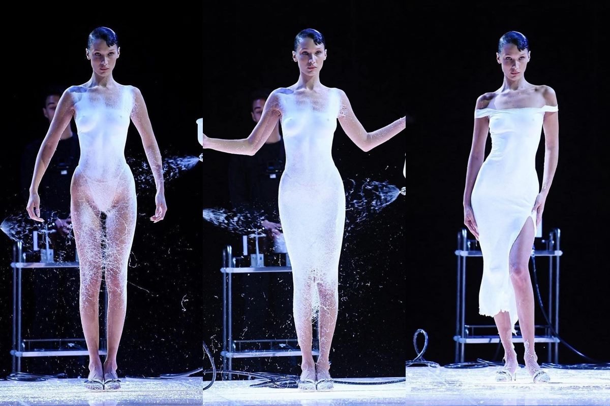 A modelo Bella Hadid, uma mulher jovem, magra, com cabelo liso preto, no desfile da marca francesa Coperni, na Semana de Moda de Paris. Ela está semi nua e uma pessoa, com um spray, borrifa uma solução que se transforma em um vestido. - Metrópoles