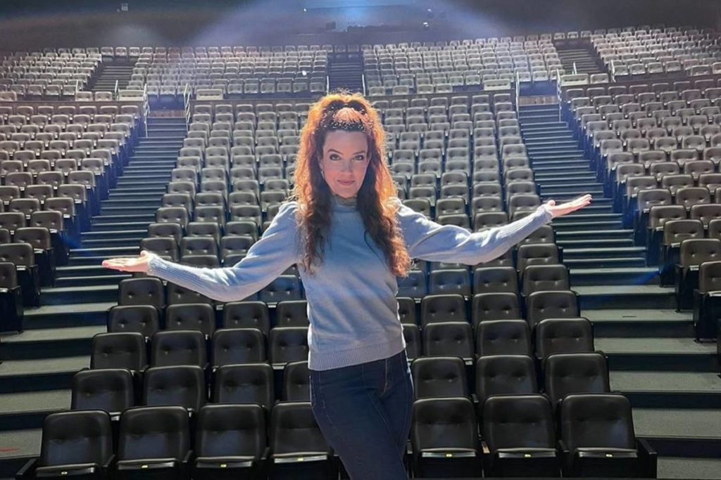 mulher de braços abertos em palco de teatro com plateia vazia ao fundo - metrópoles