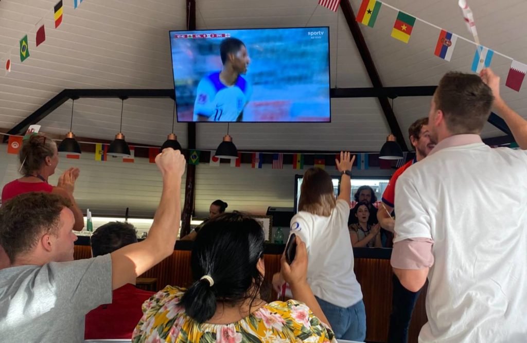 Foto de várias pessoas gritando e comemorando com as mãos para o alto o gol da Inglaterra, em último plano mostra uma TV em que mostra um jogador da Inglaterra