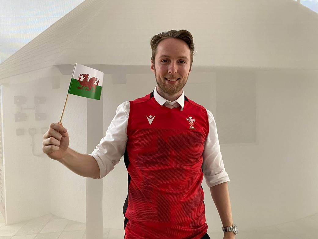 Homem branco vestindo uma blusa vermelha e segurando uma bandeira do País de Gales