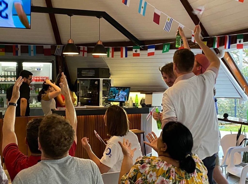 Várias pessoas em pé e com as mãos levantadas para comemorar o gol feito da Inglaterra na Copa do Mundo durante evento na Embaixada do Reino Unido