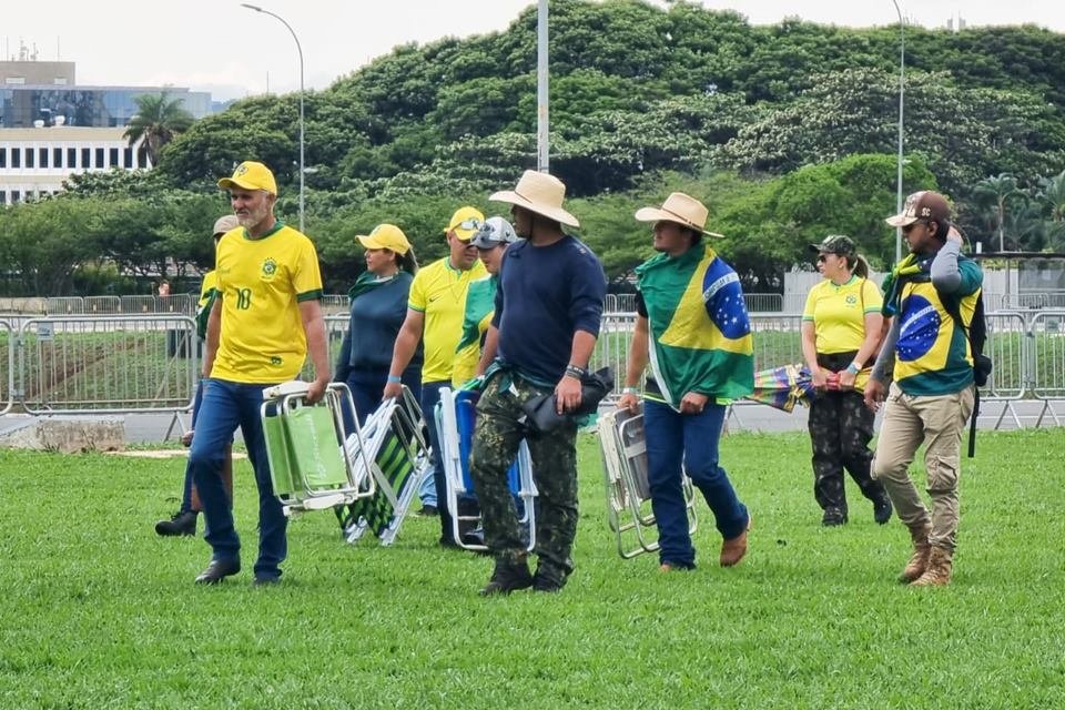 Grupo de pessoas trajadas com roupas nas cores da bandeira do Brasil e camisetas amarelas, em gramado carregando objetos como cadeiras de praia - Metrópoles