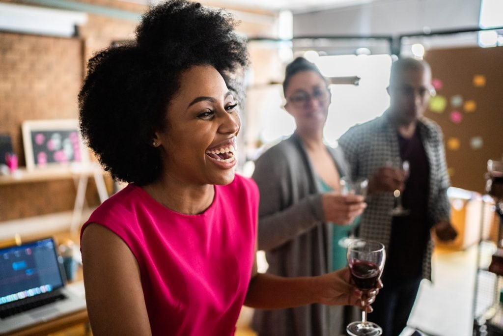 Mulher negra e jovem, de cabelo cacheado curto, fazendo um discusso durante uma confraternização do trabalho. Segura uma taça com vinho tinto e usa um blusa rosa. - Metrópoles