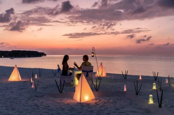 Fotografia colorida mostrando casal jantando na praia no entardecer rodeado por velas na areia-Metrópoles