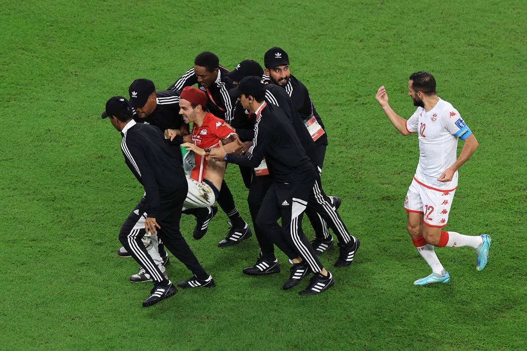 Homem com camisa e chapéu vermelhos sendo carregado por seis seguranças enquanto um jogador de roupa branco o acompanha durante invasão em jogo da Copa do Mundo